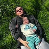 adult, baby, grass, outdoor, person, lying_down, sunglasses, clothing, baby_clothes, greenery, sunlight, casual, relaxation, father_and_child, nature, stitch, baby_socks, daylight, park, family