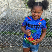 Léah joined the competition — help win amazing prizes! blue_shirt, casual_clothing, chain_link_fence, child, concrete_ground, confident, daylight, denim_shorts, earrings, fashion, girl, greenery, hair_puffs, happy, outdoor, person, portrait, smile, standing, young