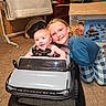 baby, box, car, car_seat, carpet, child, clothing, face, floor, furniture, hair, happy, indoor, person, play, room, sibling, smile, toy_car, vehicle