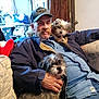 man, dog, indoor, couch, cap, mustache, denim_shirt, small_dog, scruffy_dog, pet, relaxed, seated, hand, furniture, casual_clothing, brown_hair, facial_hair, home, living_room, portrait