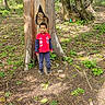 carving, child, daylight, forest, greenery, ground, leaf, nature, outdoor, owl_sculpture, pants, person, plant, red_jacket, sculpture, sneakers, standing, tree, trunk, woodland