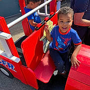 Jeremy joined the competition — help win amazing prizes! adult, blue_shirt, boy, casual_clothing, child, concrete, daytime, fire_truck, fun, happy, outdoor, person, play, playground, red, seat, smile, sunlight, toy, wood