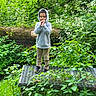 child, hoodie, standing, outdoor, greenery, plants, wildflowers, nature, forest, metal_platform, casual_clothing, young_boy, curly_hair, daylight, foliage, shoes, pants, explorer, peaceful, portrait