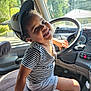 toddler, child, smiling, striped_shirt, cap, vehicle_interior, steering_wheel, sunlight, window, seat, dashboard, happy, playful, casual_clothing, hand, face, headwear, seatbelt, person, indoors