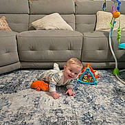 Sawyer joined the competition — help win amazing prizes! baby, child, infant, crawling, smiling, indoors, living_room, sofa, rug, baby_toys, playmat, stuffed_toy, orange_toy, activity_center, pillows, home, portrait, floor, colorful, happy