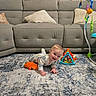 baby, child, infant, crawling, smiling, indoors, living_room, sofa, rug, baby_toys, playmat, stuffed_toy, orange_toy, activity_center, pillows, home, portrait, floor, colorful, happy