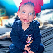 Oakley is registered to the contest to win money with this photo: baby, child, smiling, blue_eyes, red_bow, hoodie, pink_socks, sitting, bed, indoor, portrait, cute, happy, infant, young_child, clothing, person, face, headwear, cozy