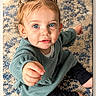 baby, barefoot, blue_eyes, brown_hair, carpet, child, close_up, home, indoor, pants, photography, pigtails, playful, portrait, reaching, rug, sitting, smiling, sweater, toddler
