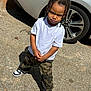 Amari is registered to the contest to win money with this photo: child, boy, braided_hair, white_tshirt, camouflage_pants, sneakers, outdoor, pavement, car, silver_car, wheel, sunlight, shadow, standing, serious_expression, young_child, street, casual_clothing, person, daytime