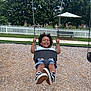 Amari is registered to the contest to win money with this photo: child, swing, playground, smiling, happy, outdoor, park, trees, fence, bench, canopy, grass, sidewalk, curly_hair, black_shoes, shorts, white_shirt, fun, summer, daytime