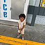 child, curly_hair, striped_clothing, shorts, sneakers, ice_machine, yellow_curb, concrete_floor, outdoor, building, door, metal_panel, standing, person, urban, casual, daylight, sidewalk, storefront, expression