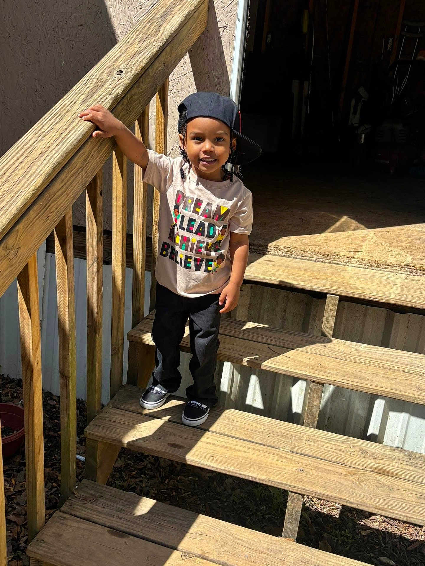 Amari is registered to the contest to win money with this photo: child, boy, standing, stairs, wooden_stairs, cap, tshirt, smile, outdoor, sunlight, shadow, handrail, casual_clothing, feet, shoes, person, daylight, happy, young, portrait