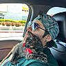 cat, tortoiseshell_cat, person, sunglasses, cap, car, passenger, road_trip, cuddle, pet, collar, red_bow, seated, portrait, indoor, sunlight, window, vehicle, fashion, companion
