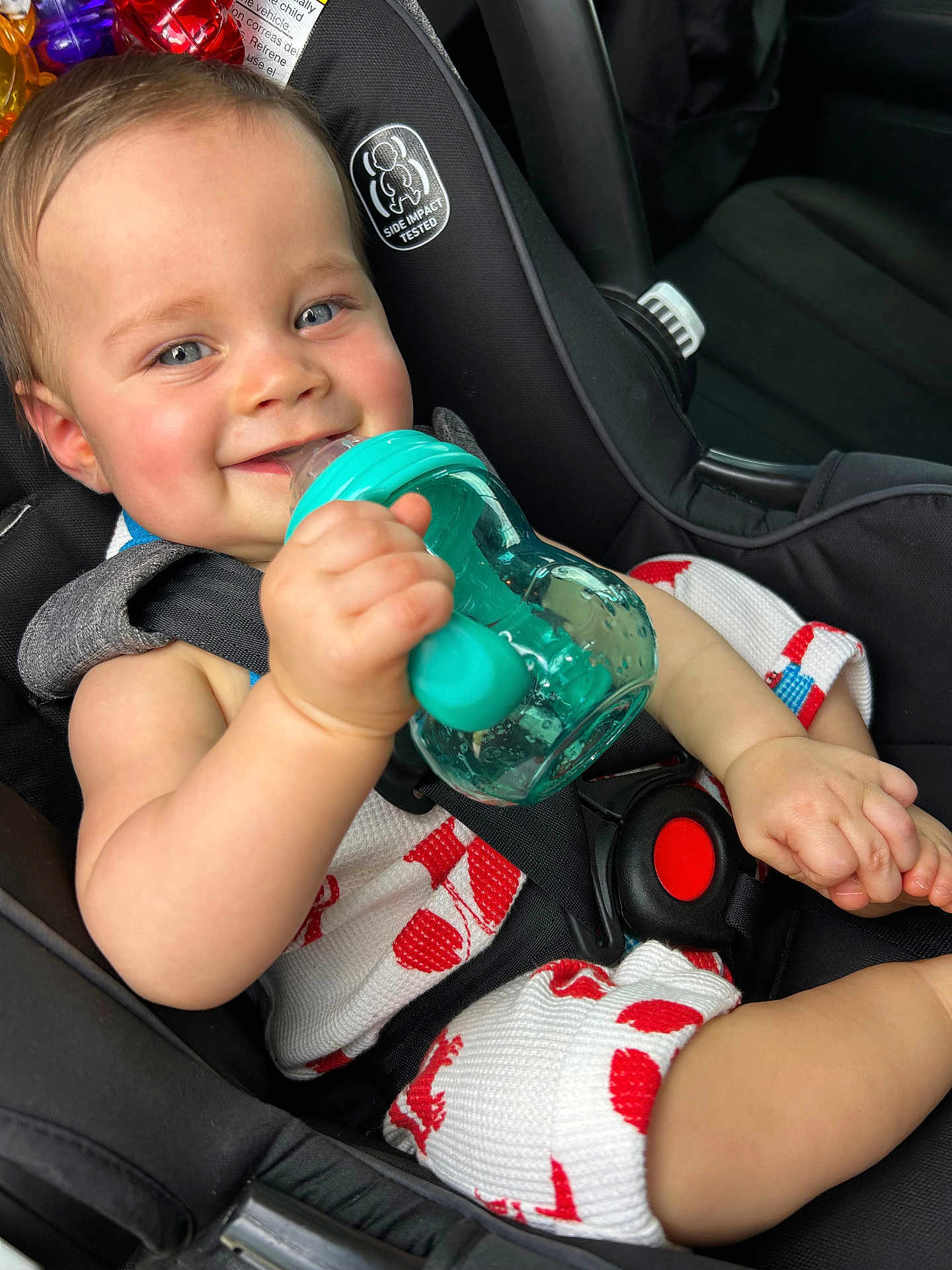 Judah is registered to the contest to win money with this photo: baby, child, car_seat, blue_eyes, smiling, sippy_cup, teal, white_outfit, red_pattern, seatbelt, infant, cute, happy, holding, hand, car_interior, portrait, closeup, comfort, safety