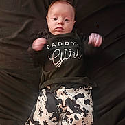 Maria is registered to the contest to win money with this photo: baby, infant, child, black_clothing, cow_print, pants, lying_down, face, hands, motion_blur, text_on_clothing, dark_background, cute, young_child, portrait, casual_clothing, fabric, indoor, person, apparel