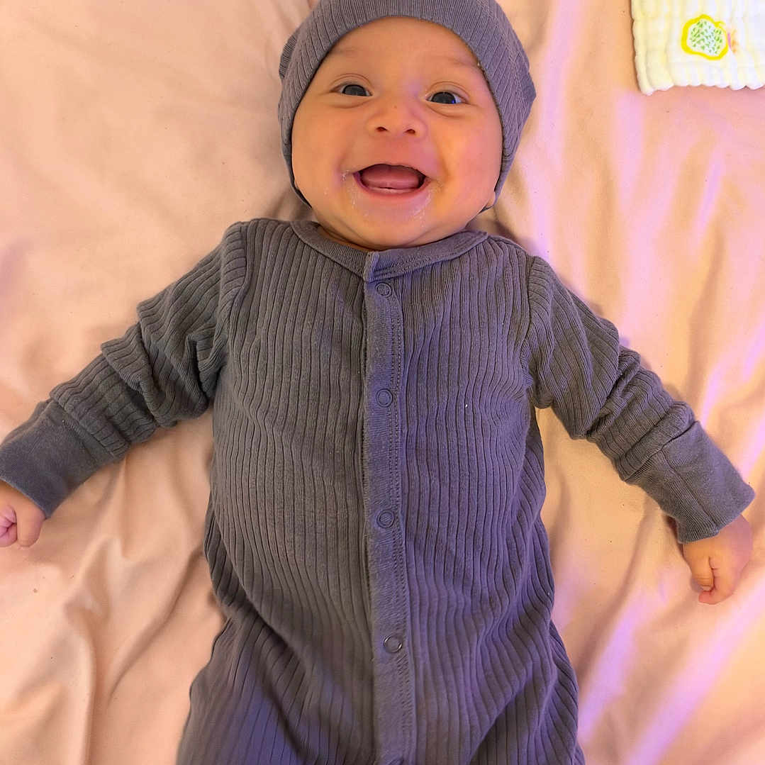 Eiden joined the competition — help win amazing prizes! baby, child, infant, smiling, happy, portrait, lying_down, bed, bedspread, gray_outfit, knit_hat, clothing, onesie, socks, indoors, home, cozy, face, photography, joy