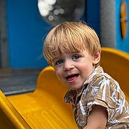 Ezekiel is registered to the contest to win money with this photo: child, toddler, blonde_hair, smiling, yellow_slide, playground, outdoor, short_sleeve_shirt, patterned_shirt, happy, face, person, summer, fun, recreation, daylight, park, cute, young, activity