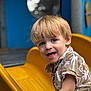 child, toddler, blonde_hair, smiling, yellow_slide, playground, outdoor, short_sleeve_shirt, patterned_shirt, happy, face, person, summer, fun, recreation, daylight, park, cute, young, activity