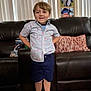 child, boy, smiling, barefoot, shirt, shorts, flamingo_pattern, couch, leather, pillow, indoor, carpet, window_blinds, photograph, stacked_clothes, portrait, happy, home, furniture, person
