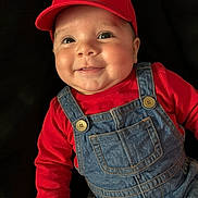 Greyson is registered to the contest to win money with this photo: baby, child, smiling, red_cap, denim_overalls, red_shirt, face, portrait, cute, indoor, infant, happy, fashion, clothing, person, headwear, button, closeup, cheeks, playful