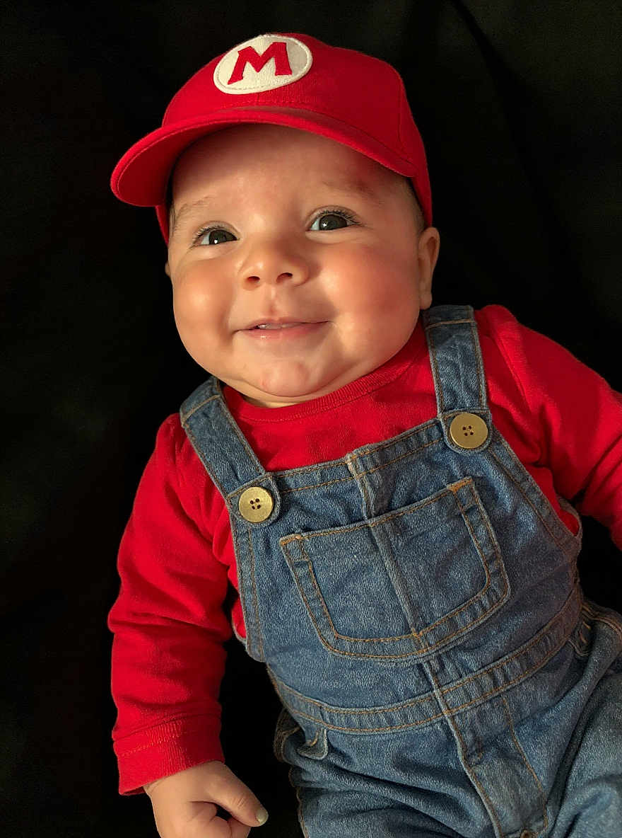 Greyson is registered to the contest to win money with this photo: baby, child, smiling, red_cap, denim_overalls, red_shirt, face, portrait, cute, indoor, infant, happy, fashion, clothing, person, headwear, button, closeup, cheeks, playful