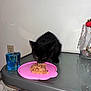 black_kitten, cat, wet_food, pink_plate, glass_table, blue_glass, jar, crystals, roller, plastic_bottle, floor, white_wall, indoor, pet, animal, feline, eating, tabletop, small, cute