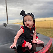 Draven is registered to the contest to win money with this photo: baby, child, car, black_car, hooded_outfit, mouse_ears, blue_eyes, outdoor, field, cloudy_sky, reflection, sitting, cute, infant, red_trim, vehicle, portrait, curious, barefoot, nature