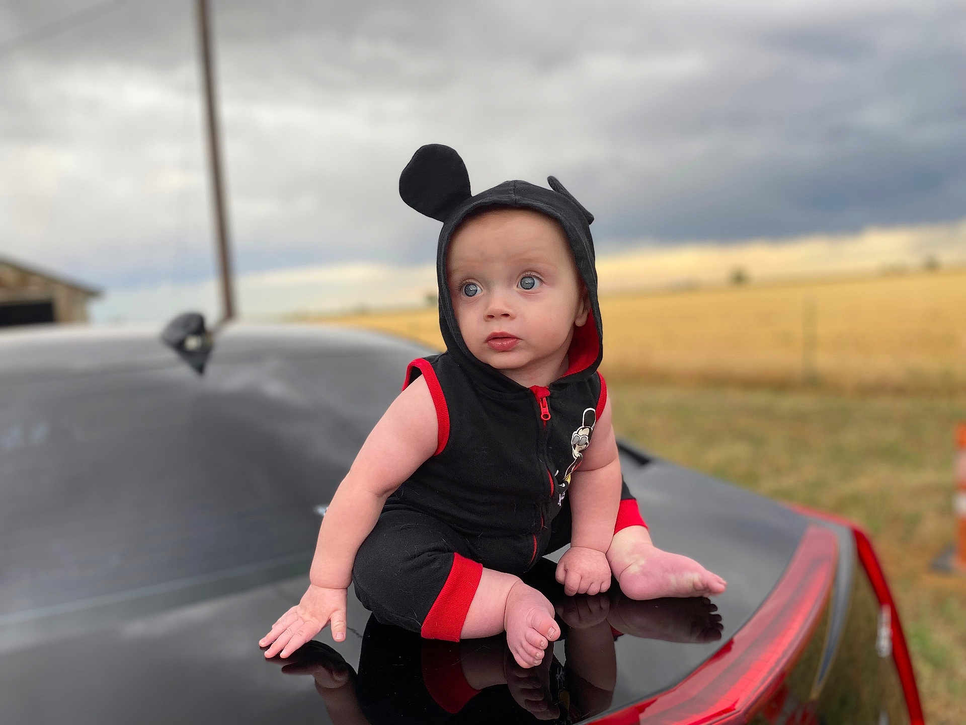 Draven is registered to the contest to win money with this photo: baby, child, car, black_car, hooded_outfit, mouse_ears, blue_eyes, outdoor, field, cloudy_sky, reflection, sitting, cute, infant, red_trim, vehicle, portrait, curious, barefoot, nature