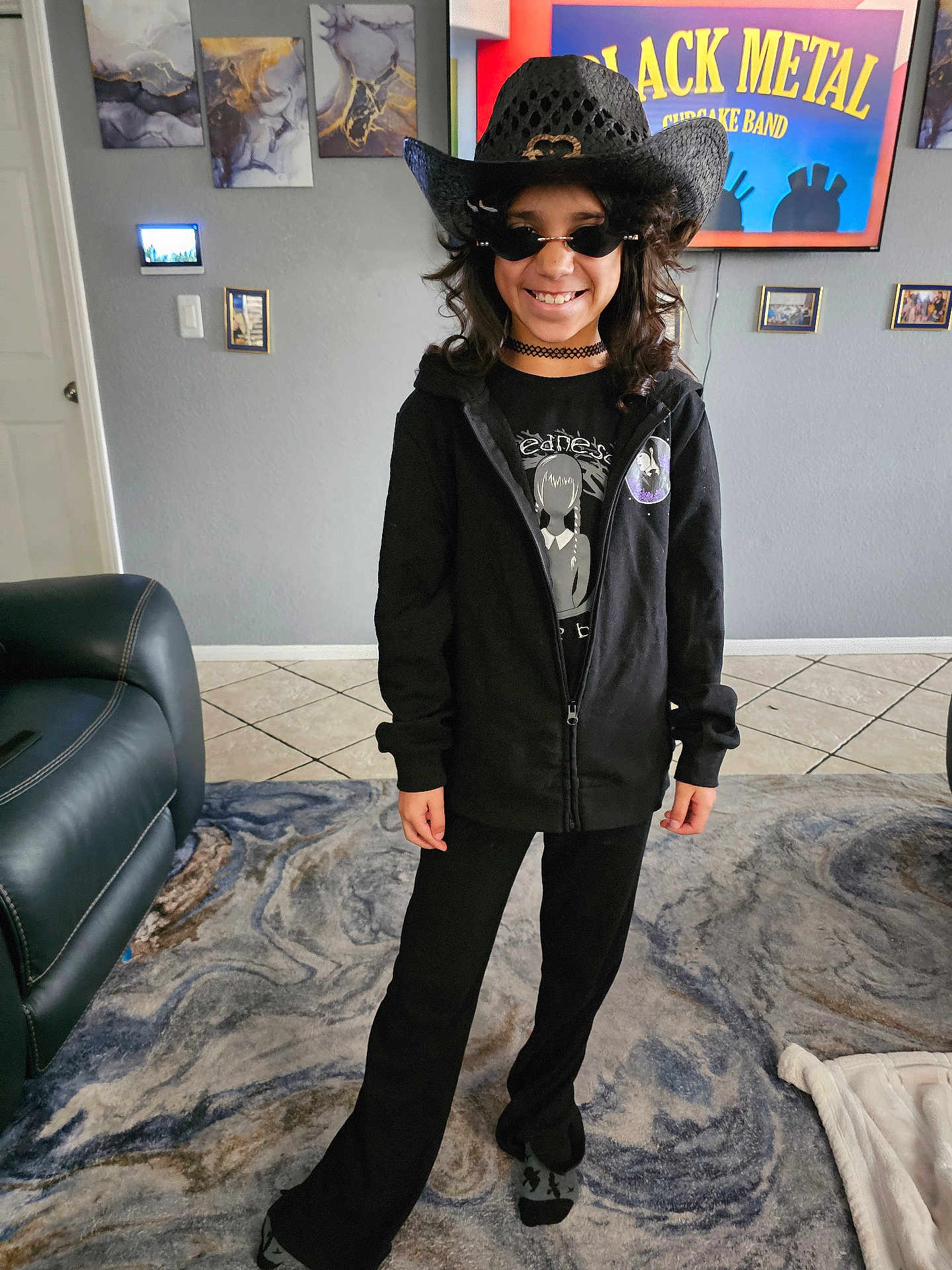 Avianna is registered to the contest to win money with this photo: child, indoor, black_clothing, cowboy_hat, sunglasses, smiling, standing, rug, living_room, sofa, tile_floor, wall_art, television, long_hair, choker, casual, person, home_decor, black_metal, happy