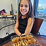 Avianna is registered to the contest to win money with this photo: child, girl, smiling, sushi, food, table, indoor, window, car, container, black_clothing, long_hair, cheerful, seat, chair, wooden_table, plastic_container, daylight, restaurant, casual