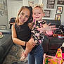 baby, black_top, carpet, child, couch, family, floral_outfit, happy, headband, holding, home, indoor, jeans, living_room, person, photograph, siblings, smiling, toy, wall_art