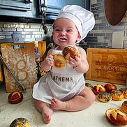 Athena is registered to the contest to win money with this photo: apron, baby, baking, bread, chef_hat, child, cooking, countertop, cute, flour, food, happy, infant, kitchen, messy, pastry, playful, rolls, sitting, smiling