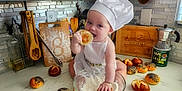 Athena is registered to the contest to win money with this photo: apron, baby, baking, bowl, bread_roll, cafe, chef_hat, child, cooking, countertop, cutting_board, flour, food, indoor, kitchen, person, playful, smiling, toddler, wooden_utensils