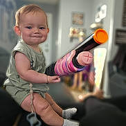 Athena joined the competition — help win amazing prizes! baby, child, toy_gun, indoor, sitting, smiling, cute, socks, pacifier, green_clothing, face, person, home, furniture, blurred_background, living_room, infant, happy, playful, short_hair
