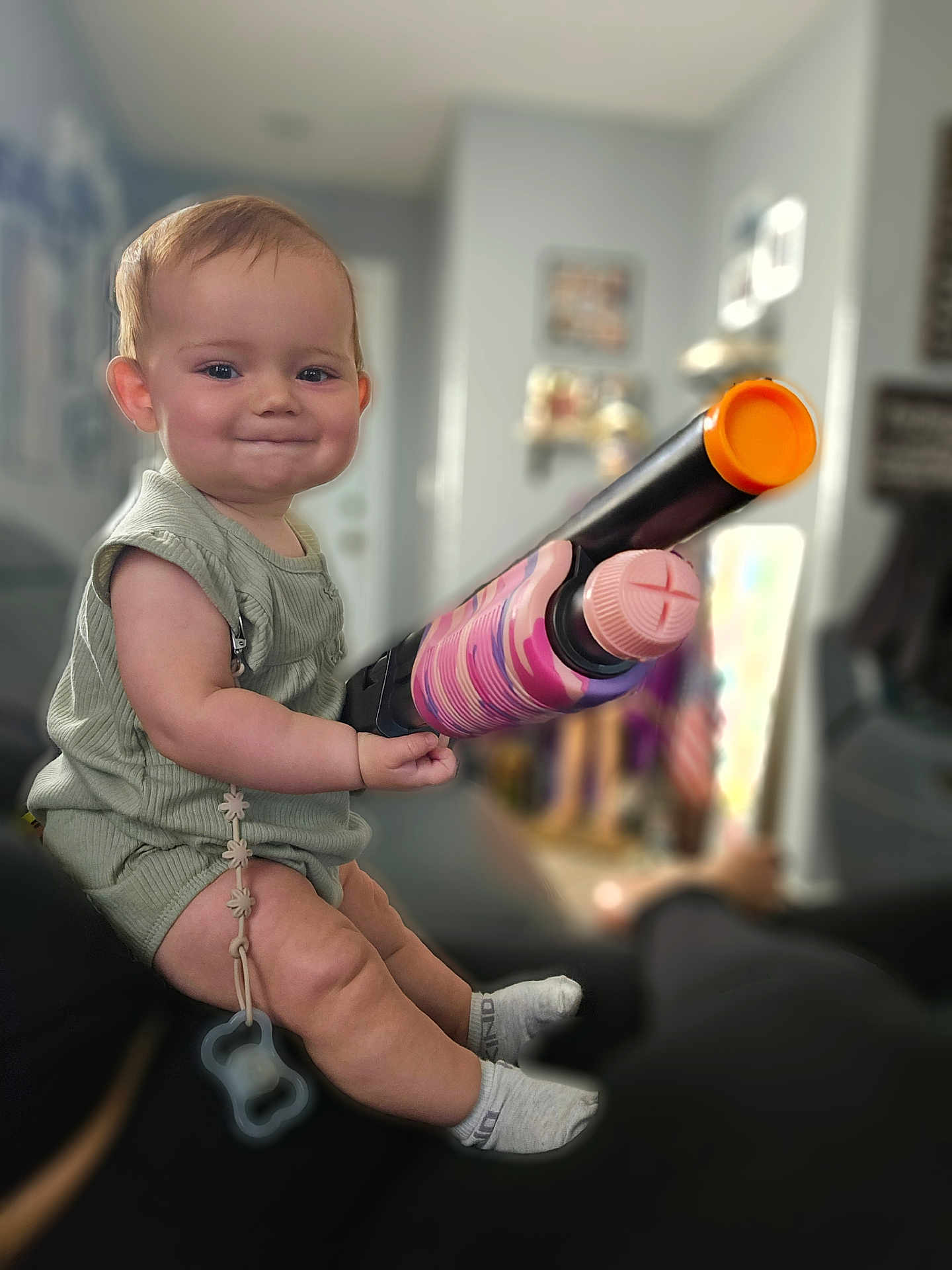 Athena joined the competition — help win amazing prizes! baby, child, toy_gun, indoor, sitting, smiling, cute, socks, pacifier, green_clothing, face, person, home, furniture, blurred_background, living_room, infant, happy, playful, short_hair