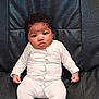 baby, infant, onesie, white_clothing, black_leather, couch, furniture, indoor, child, person, face, curly_hair, sitting, expression, cute, young, portrait, comfortable, home, serious