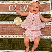 Olivia is registered to the contest to win money with this photo: baby, child, infant, smiling, pink_clothing, headband, bow, crochet_blanket, wooden_plaque, three_months, happy, lying_down, face, cute, skin, floor, patterned_rug, decor, portrait, indoors