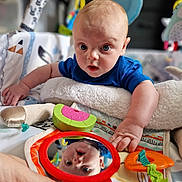 Eli joined the competition — help win amazing prizes! baby, infant, mirror, reflection, blue_shirt, toy, hand, blanket, play_area, colorful, face, expression, indoor, child, curious, soft_toy, closeup, cute, person, reflection_image