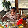 baby, infant, child, headband, shirt, baby_phat, activity_center, toy, table, chair, plant, window, blinds, indoor, smiling, cute, person, furniture, wood, happy