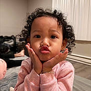 Christina is registered to the contest to win money with this photo: child, toddler, portrait, curly_hair, kissy_face, pouting, hands_on_cheeks, smiling, pink_shirt, long_sleeves, bedroom, bed, indoor, curtains, soft_lighting, close_up, sitting, cozy, adorable, photography