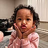 child, toddler, portrait, curly_hair, kissy_face, pouting, hands_on_cheeks, smiling, pink_shirt, long_sleeves, bedroom, bed, indoor, curtains, soft_lighting, close_up, sitting, cozy, adorable, photography