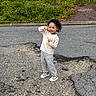 child, toddler, young_girl, portrait, smiling, playful, peace_sign, hand_gesture, curly_hair, long_sleeves, striped_pants, sneakers, outdoor, road, asphalt, cracked_pavement, greenery, hedge, daylight, full_body