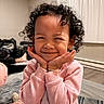 child, toddler, smiling, happy, curly_hair, portrait, indoor, bedroom, bed, blanket, pink_clothing, floral_pants, hands_on_cheeks, sitting, playful, cute, curtains, window, home, photography