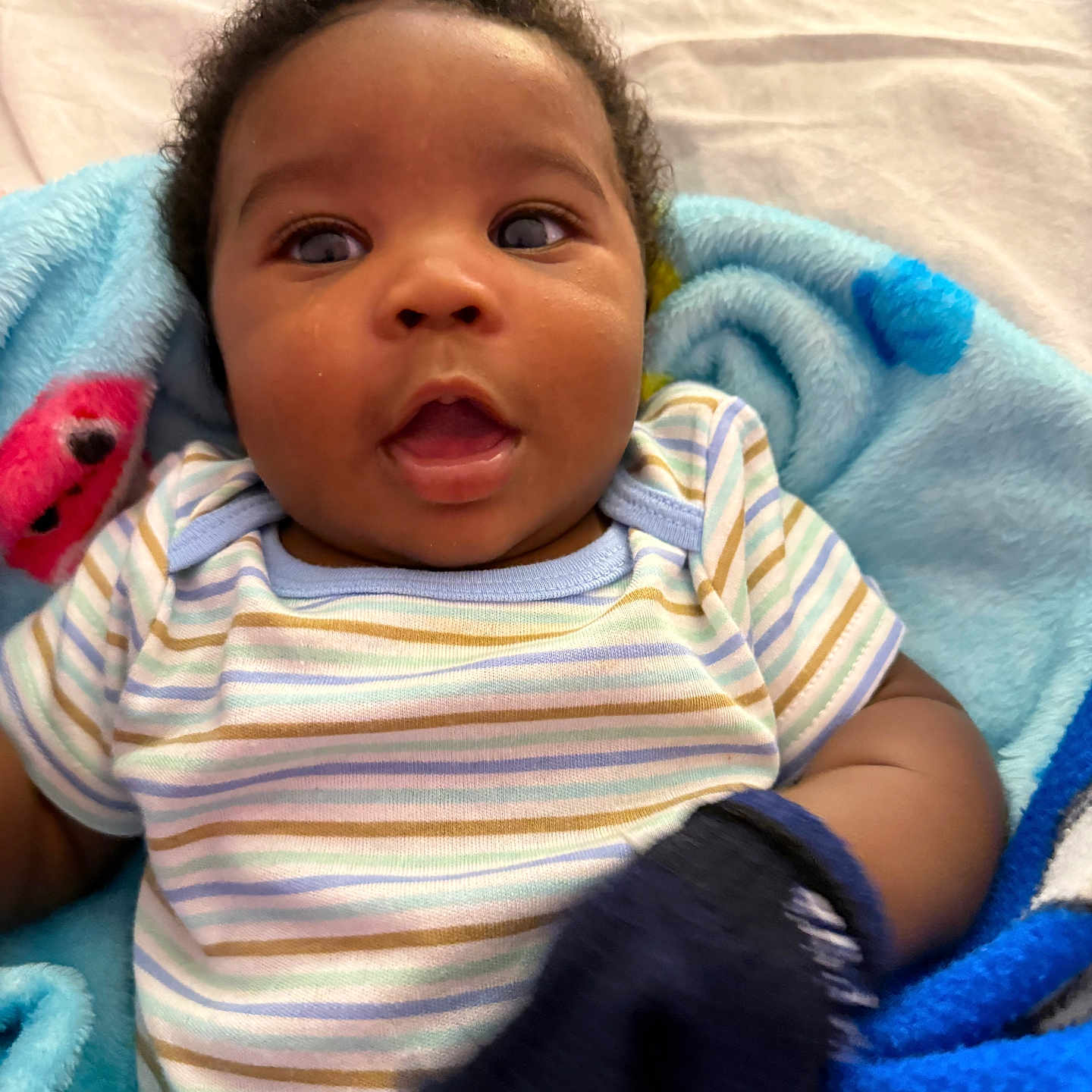 Jermany is registered to the contest to win money with this photo: baby, infant, child, blanket, striped_clothing, mitten, face, curly_hair, expression, portrait, indoors, cozy, happy, cute, soft_texture, blue, multicolor, closeup, person, newborn