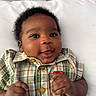 Jermany is registered to the contest to win money with this photo: baby, infant, child, smiling, face, plaid_shirt, clothing, hand, lying_down, bed, pillow, bottle, background, cute, portrait, skin, hair, eyes, happy, indoors