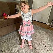 Zoey is registered to the contest to win money with this photo: child, girl, smiling, dress, pink_bow, living_room, carpet, couch, sippy_cup, home_interior, wood_floor, door, lamp, pillow, happy, person, footwear, sandals, wall_clock, curtains