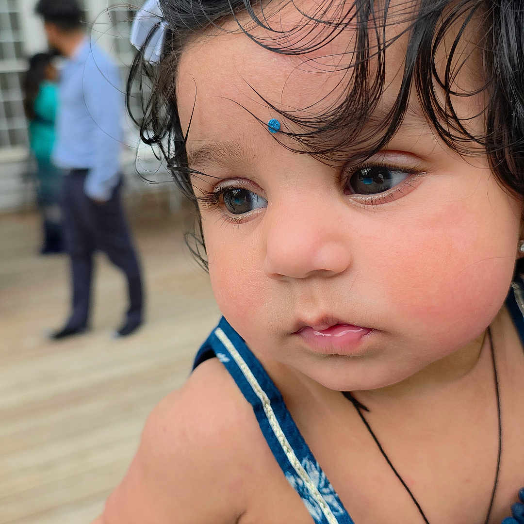 Aaira is registered to the contest to win money with this photo: toddler, child, face, curly_hair, blue_bindi, blue_dress, outdoor, wooden_deck, person, hand, portrait, cute, skin, expression, young_child, holding, background_people, daylight, closeup, thoughtful