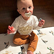Dutton is registered to the contest to win money with this photo: baby, child, pacifier, bed, duck_pattern, white_shirt, brown_pants, sitting, curious_eyes, indoors, soft_light, cute, infant, foot, hand, face, young_child, clothing, person, portrait