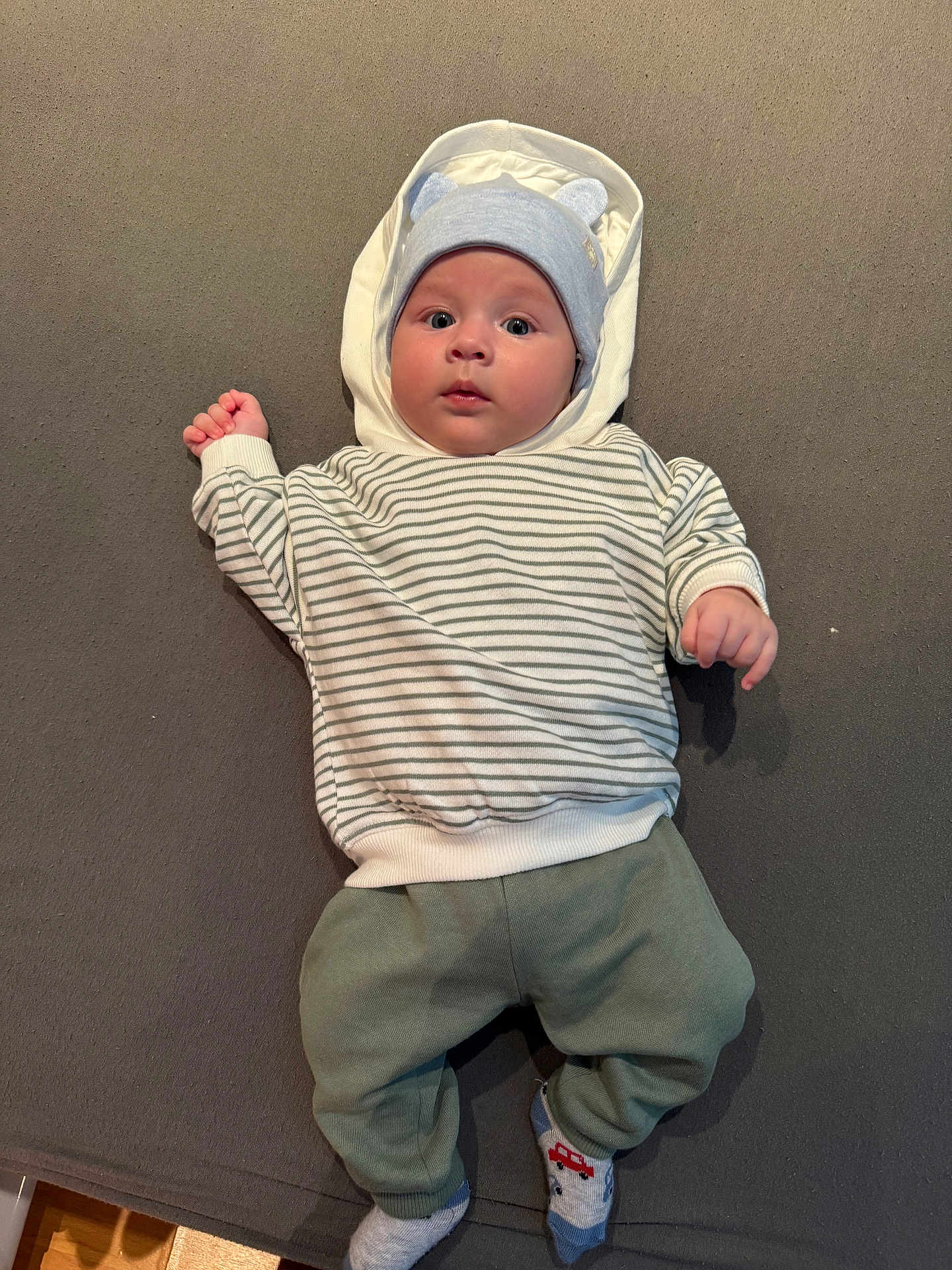 Thomas a rejoint le concours — aidez-le/la à gagner de superbes lots ! baby, infant, hoodie, striped_sweater, blue_hat, socks, car_pattern, lying_down, grey_background, cute, child, portrait, clothing, casual, indoor, small_hands, face, headwear, comfort, soft_surface