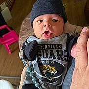 Leonidas is registered to the contest to win money with this photo: baby, infant, clothing, hat, person, indoor, holding, hand, face, expression, curious, black_hat, jaguars, football, pacifier_clip, wooden_floor, pink_chair, plant, tattoo, casual